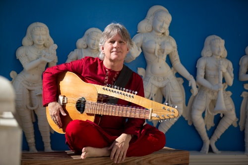 man sitting cross-legged in front of Indian statues holding a combination guitar/sitar instrument