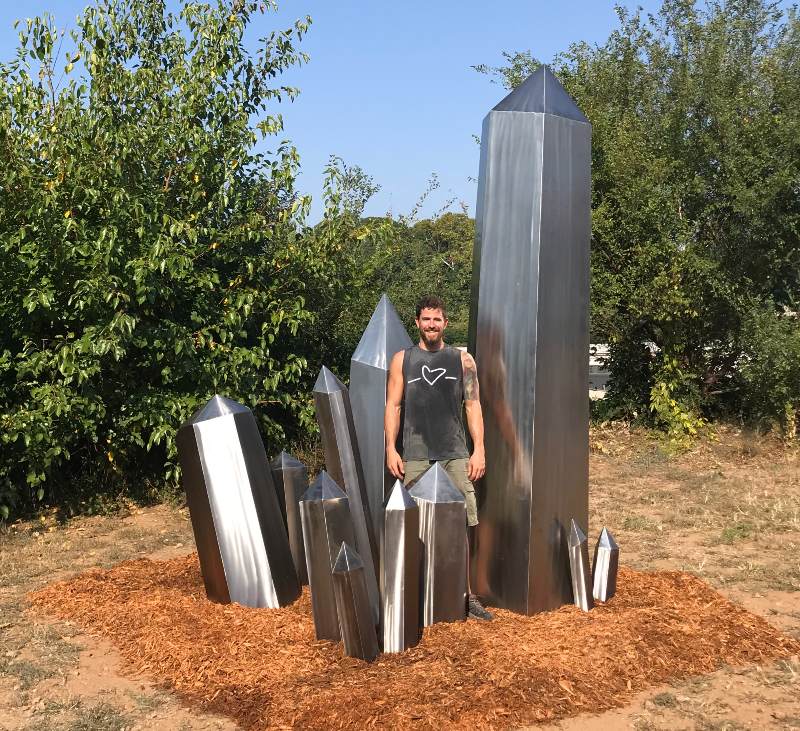 Man standing within sculpture consisting of many shiny silvery obelisks close together, one twice as high as he is, one his height, several smaller, set on a bed of mulch with large green bushes behind it