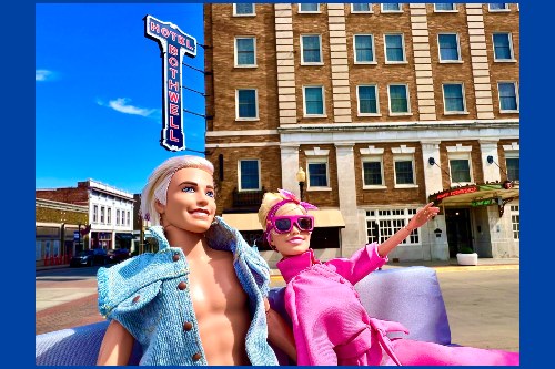 Photo of Barbie and Ken dolls, her in Barbie pink, him in blue denim, sitting on satin couch with old downtown hotel building in the background, with perspective as though Barbie and Ken were as large as real people