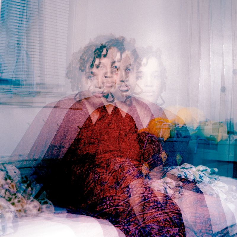 Photo of woman in red dress sitting on the edge of a twin bed, with multiple exposures so that she is quadrupled with each of her images slightly overlaps the others