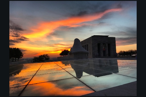 Photo of massive building in classical style, in distance, seen across large platform of mirrored tiles in which a flaming sunset is dramatically reflected