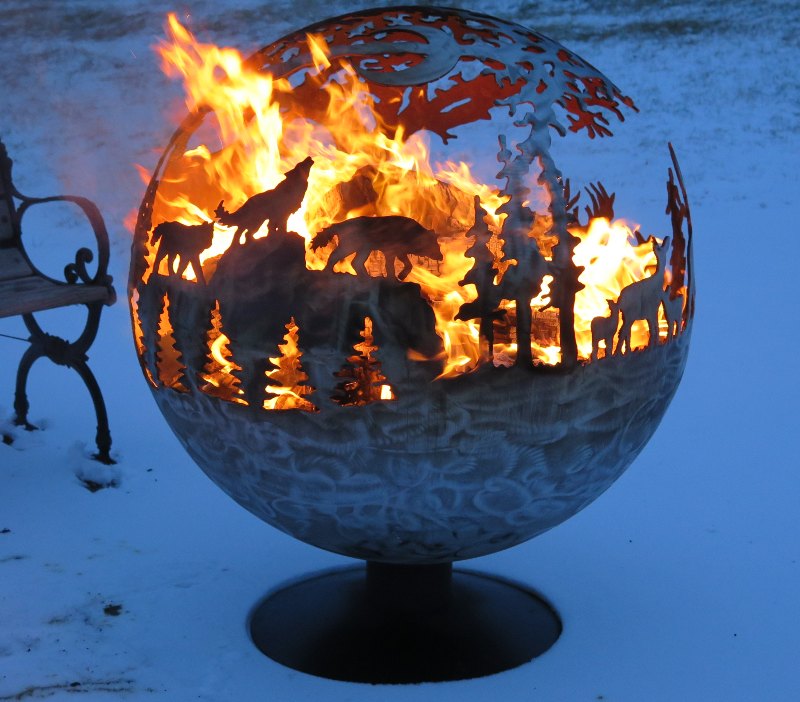 Large metal globe outdoors in the snow, bottom solid, top carved with silhouettes of pine trees, howling wolves, doe with two fawns, and starry sky overhead, backlit by flames blazing inside the globe