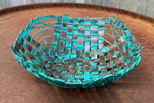 Shallow bowl woven in large strips of metal in shades from bright copper to turquoise blue