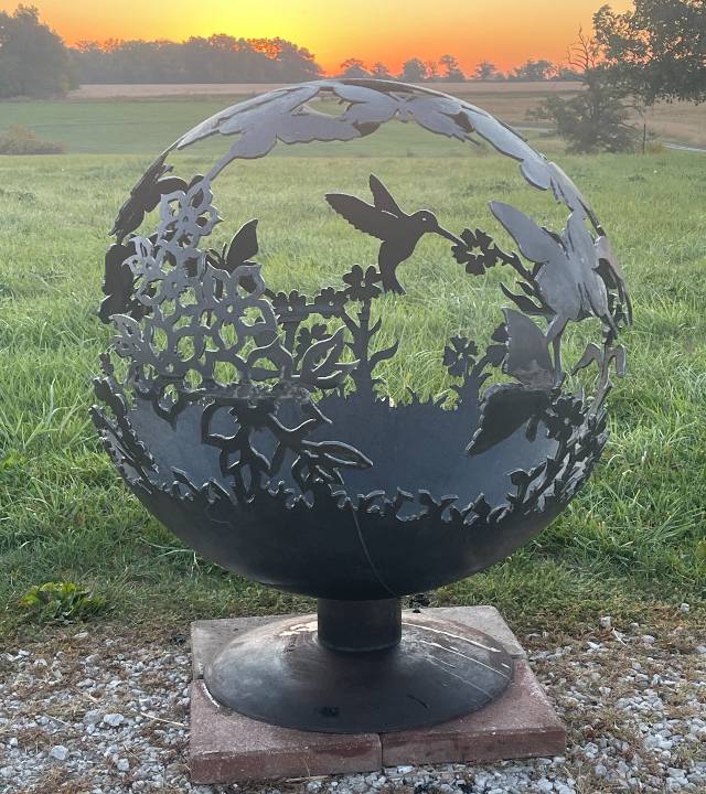 Large metal globe outdoors in a field, bottom solid, top carved with silhouettes of butterflies, hummingbirds, and flowers