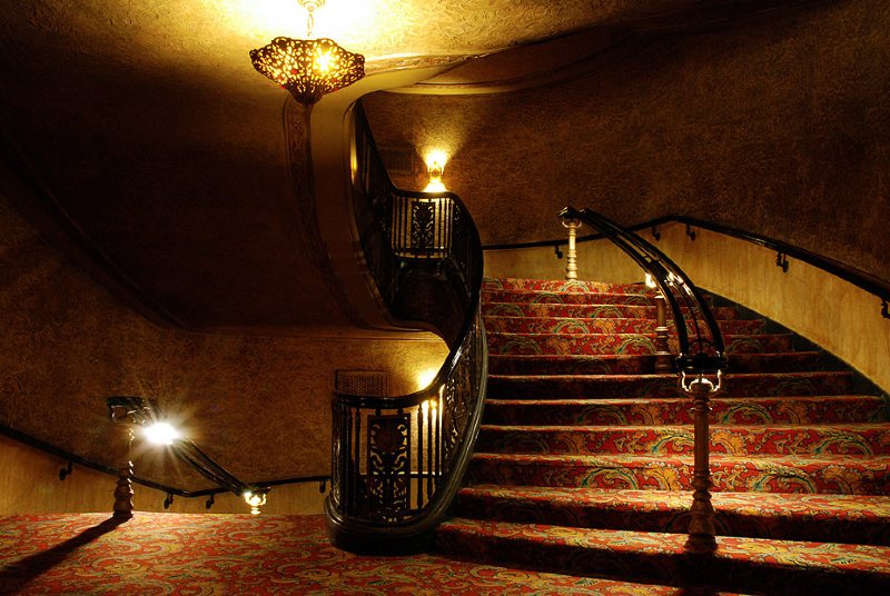 Photograph of stairway landing and stair leading upwards, gold walls, richly patterend carpet in reds and golds, antique lamps casting gold light and deep shadows