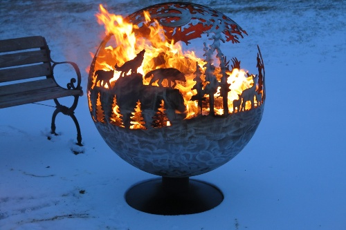 Large metal globe outdoors in the snow, bottom solid, top carved with silhouettes of pine trees, howling wolves, doe with two fawns, and starry sky overhead, backlit by flames blazing inside the globe