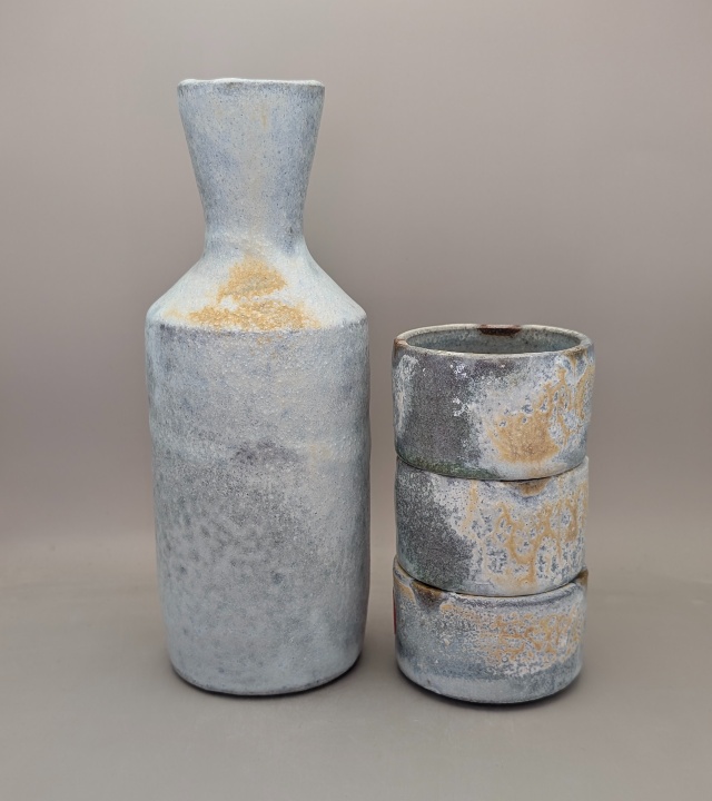 Photo of bottle next to three small cups without handles stacked up, rough-textured finish in shades of light blue and tan