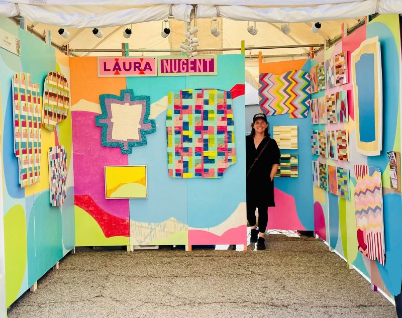 Inside of artist's booth, three walls covered with colorful abstract geometric paintings, with smiling artist standing at back of booth