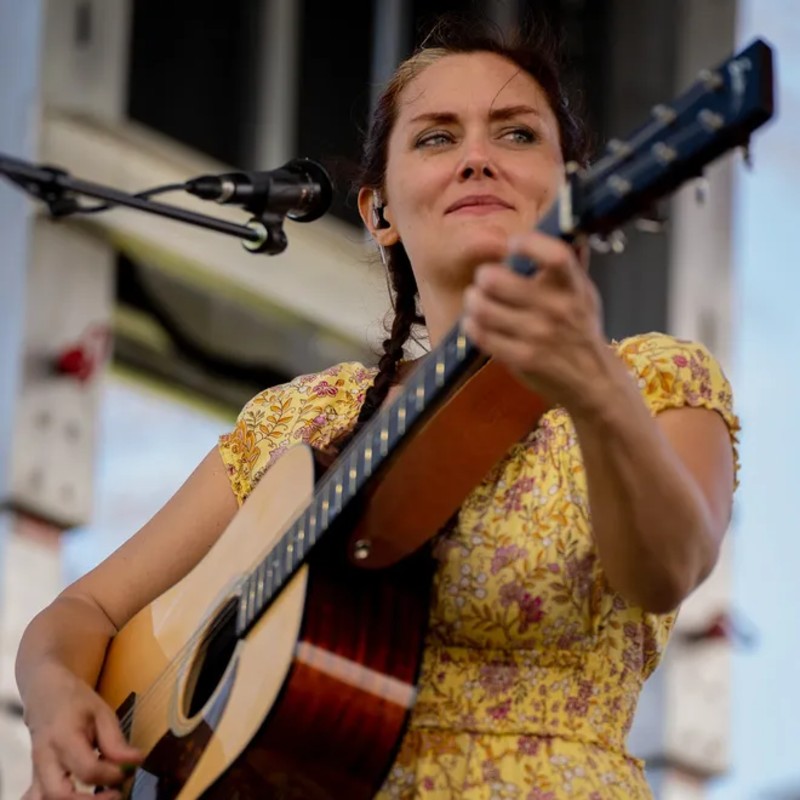 Woman in yellow floral dress outdoors standing in front of a microphone and playing a guitar