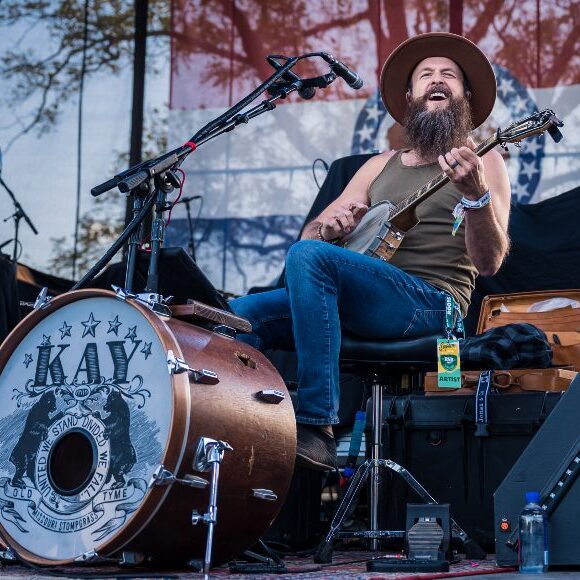Man seated on outdoor stage singing and playing a banjo, wearing jeans, A-shirt, and cowboy hat
