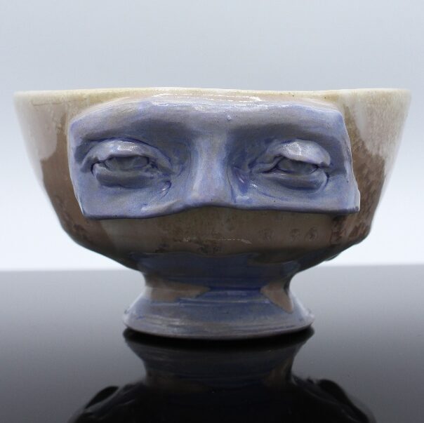 Footed ceramic bowl with sculpture on the side of partial face, nose and two eyes, in muted blues and creams