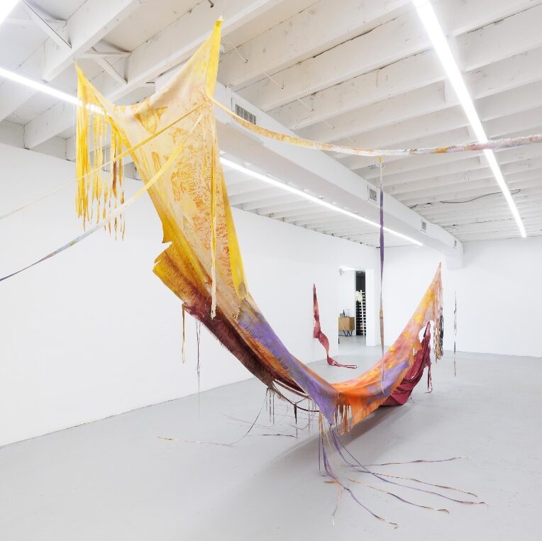 View of a large high-ceilinged room with white walls, large fabric sculpture filling the middle, a long swath with both ends fastened to rise toward the ceiling