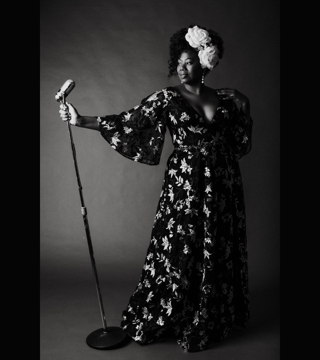 Dramatically lit black-and-white full-length portrait photo of woman standing at microphone, wearing elegant long dress and large roses in her hair