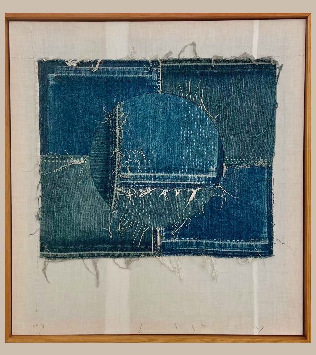 Framed artwork of patches of denim, stitched together and over-stitched, mounted on cream-colored plain fabric