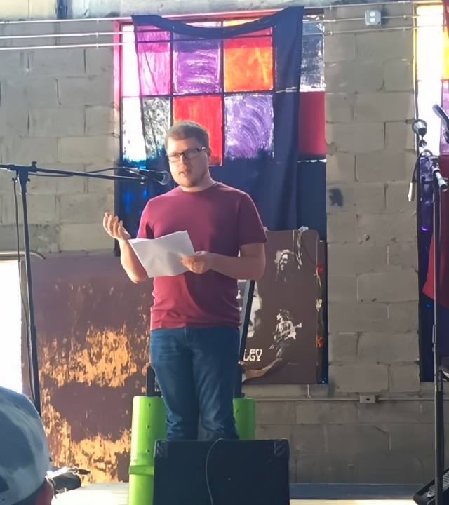 Man performing his poetry, standing at microphone on interior stage holding papers