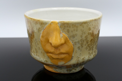 Footed ceramic bowl with sculpture on the side of partial face, nose and lips, in muted golds and greens