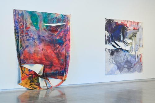 Wall in gallery with two large flat fabric sculptures, multi-colored abstract patterns, rips in the fabric