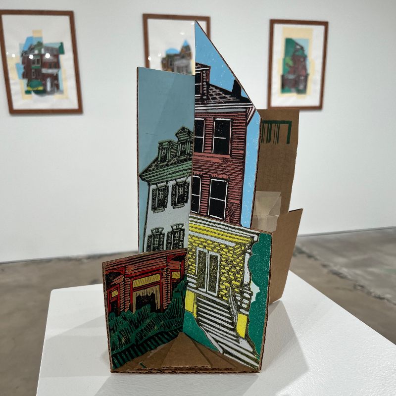 Photo of table at art exhibit with three-dimensional collage of drawings of various pieces of exteriors of different houses mounted on cardboard and assembled into a "combination house"