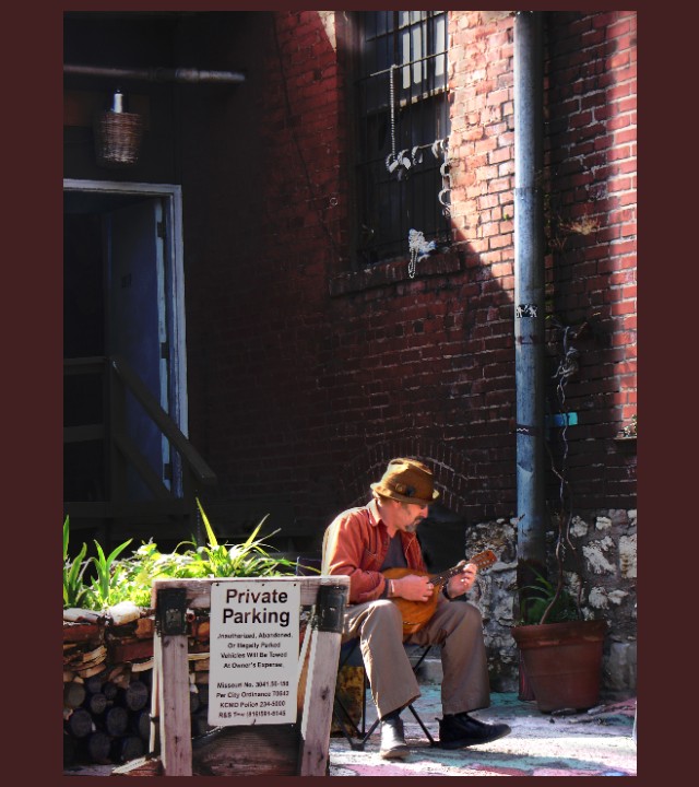 Photograph of a nook on a city sidewalk formed by two brick walls at right angles with a man seated between them playing a guitar