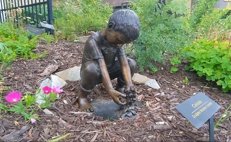 Statue in bronze of a young child crouching down to touch flowers emerging the earth, in the setting of a small garden with low-lying green and flowering plants