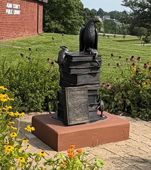 Statue in bronze of a stack of books, the bottom book with the letters GATHER, with a raven on top and a rabbit, frog, and turtle climbing the stack; situated on a terra cotta-colored base in a garden outside a red brick building