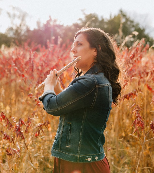 Photograph of a woman standing in a field of tall red and gold plants, playing a flute