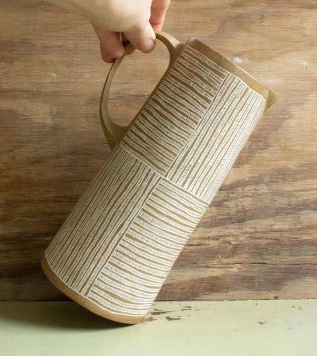 Pottery pitcher in the shape of a simple tall narrow cylinder, tan color patterned with bands of white lines