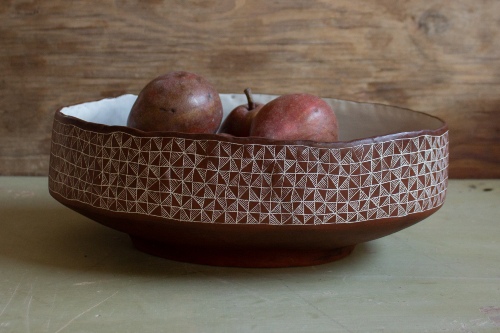 Oval bowl seen from its side, red color patterned with squares and triangles filled with white lines, with three red pears inside the bowl