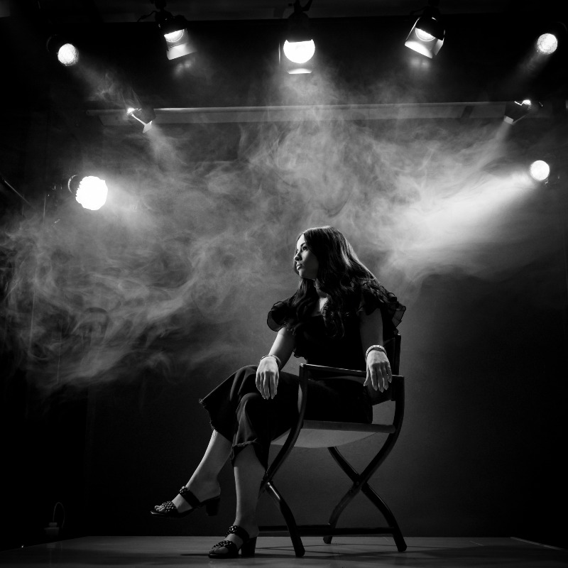 Dramatically lit black and white photograph of woman sitting on chair underneath seven floodlights with illuminated fog swirling above her head