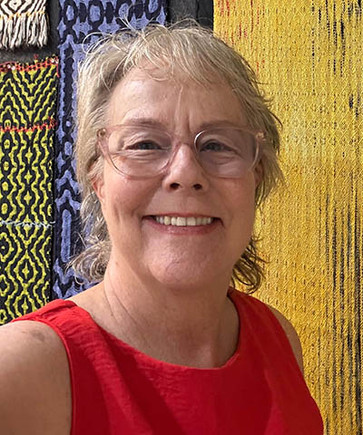 head-and-shoulders portrait, older woman with glasses in red shirt against multi-colored background of woven textiles